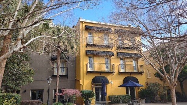 A yellow building with balconies, a covered patio area with outdoor seating, and surrounding greenery.