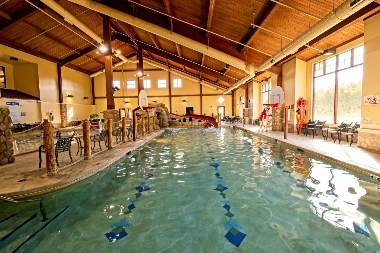 Une piscine intérieure avec un plafond en bois, un plongeoir et un espace détente avec des tables et des chaises.