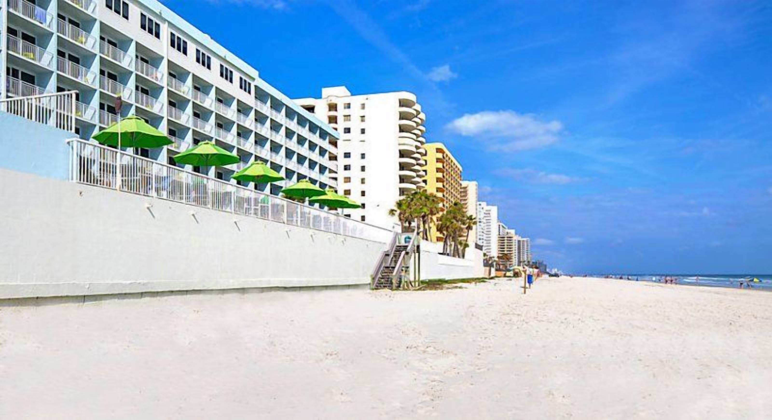 Un hotel frente a la playa con varios balcones, una playa de arena y un cielo azul despejado.