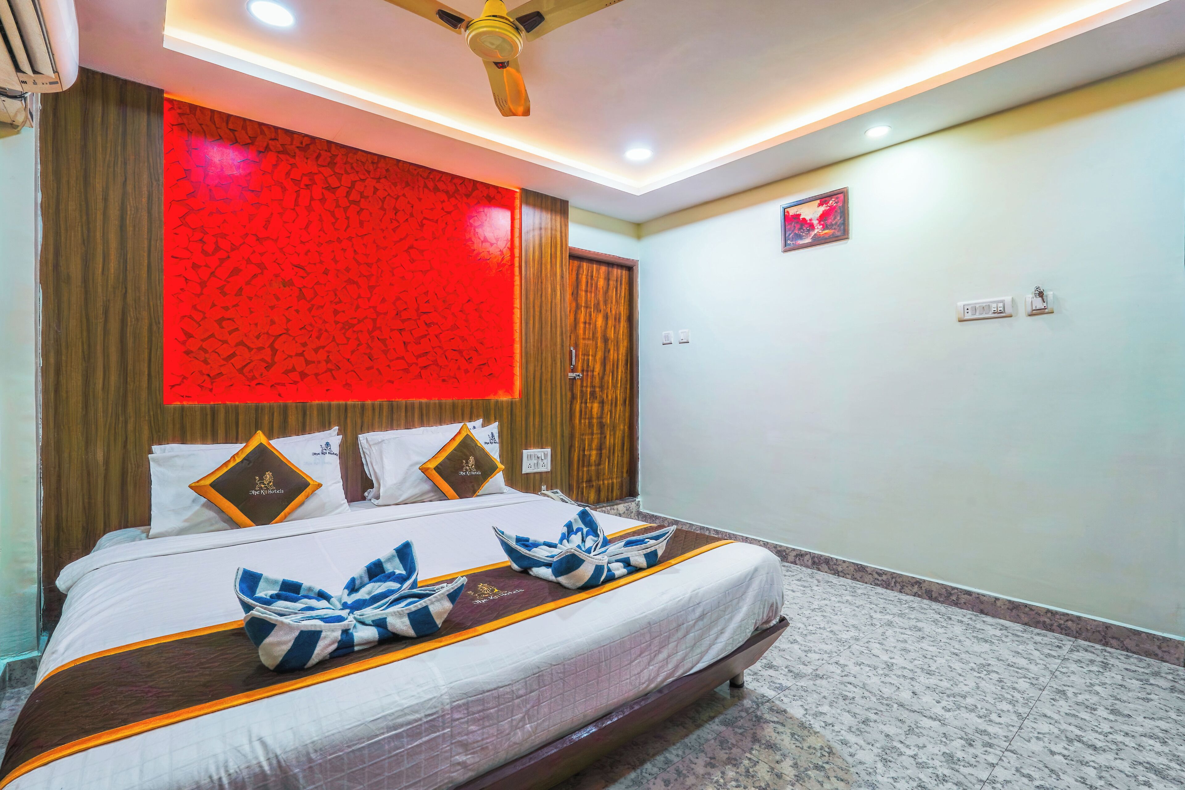 A hotel room with a bed, a ceiling fan, a red decorative wall, and a framed picture on the wall.