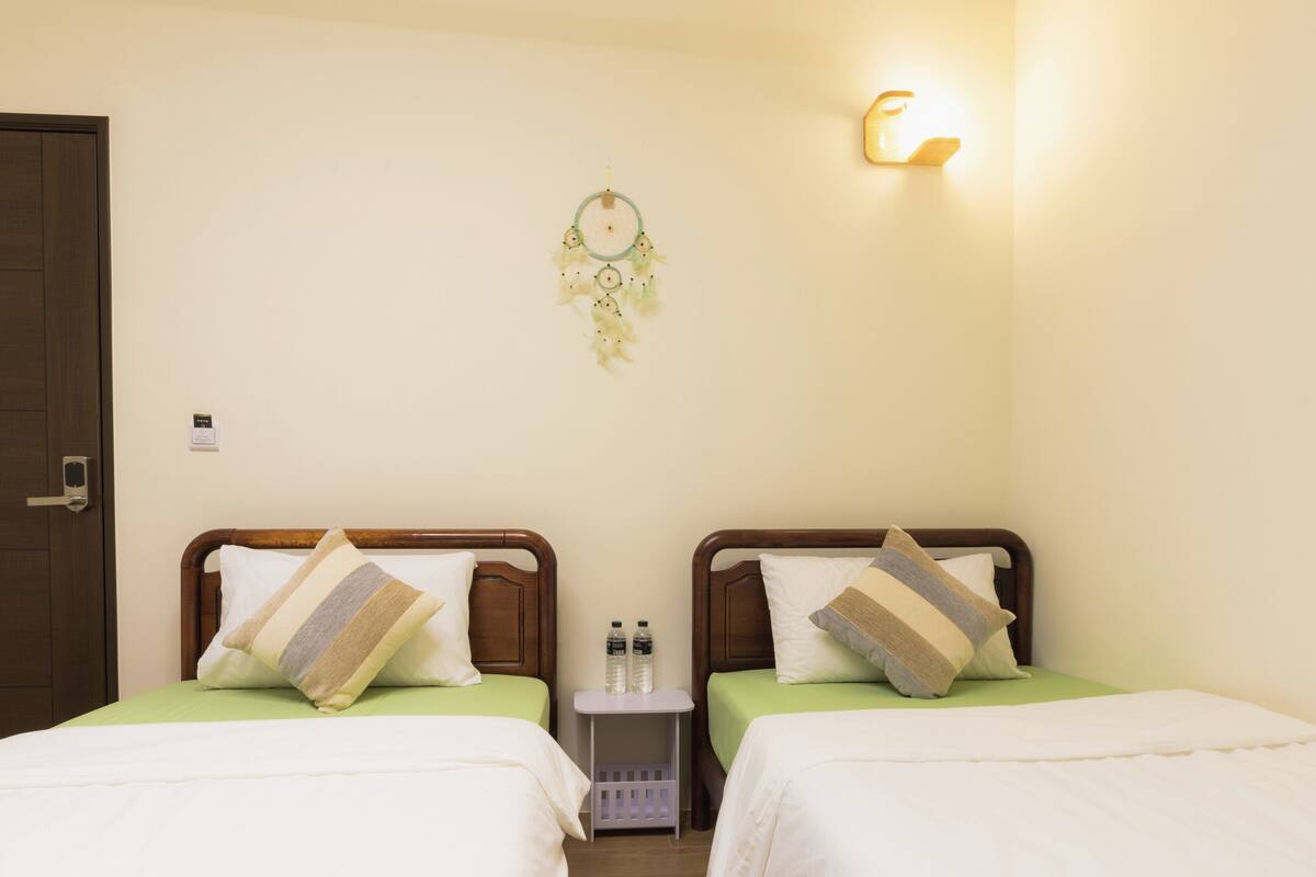 A hotel room with two beds, each with a wooden headboard and a pillow. There is a bedside table with a water bottle and a small plant. A dreamcatcher hangs on the wall, and a light fixture is mounted above the beds.