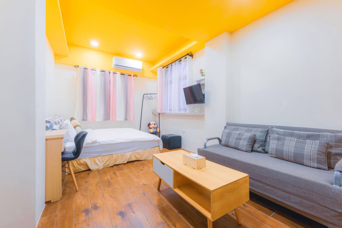 A compact room with a bed, a desk, a sofa, and a TV mounted on the wall.
