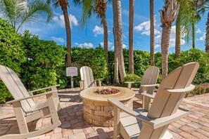 A patio area with Adirondack chairs and a stone fire pit surrounded by lush greenery and palm trees. - Orlando | Family Theme Park Retreat | Pool + Hot Tub | 2BR (Orlando)