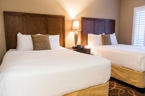 A hotel room with two beds, a nightstand with a lamp, and a window with blinds.