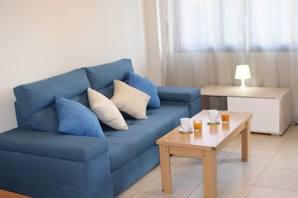 A blue sofa with cushions, a wooden side table with a lamp, and a coffee table with cups and a glass.