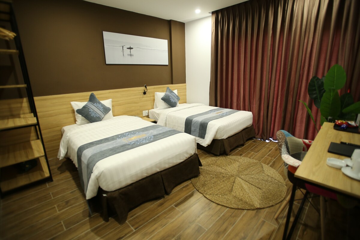 A hotel room with two beds, a wooden headboard, a wall-mounted artwork, a round woven rug, a desk with a chair, and a large window with curtains.