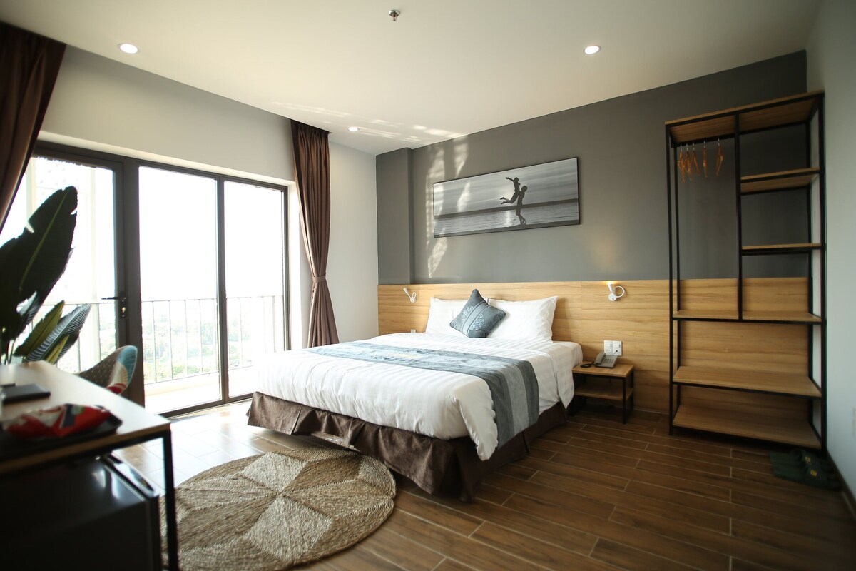 A modern bedroom with a large bed, a wooden headboard, a bedside table, and a view of the outdoors.