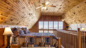 Une chambre à coucher aménagée dans une cabane en bois, avec un lit, une table de chevet, une lampe, un ventilateur de plafond et une vue sur les montagnes grâce à de grandes fenêtres.