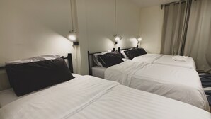 A hotel room with three single beds, each with white bedding and black pillows. There are two bedside lamps on each bed. The room has a simple, functional design with plain walls and a window with curtains. - House 116 (Bangkok)