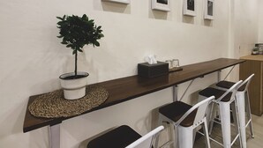 A long, dark wooden table with a woven placemat, flanked by white barstools. A potted plant is on the left side of the table. - House 116 (Bangkok)