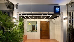 A modern hotel entrance with a wooden door, a digital display showing room numbers, and potted plants. - House 116 (Bangkok)
