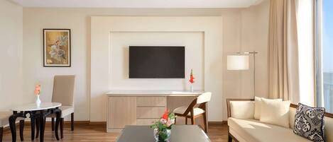 A modern hotel room with a sofa, a dining table, a TV mounted on the wall, and a large window with curtains.