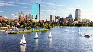 A city skyline with modern buildings, a river with sailboats, and a park with trees.