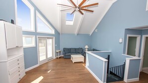 A spacious living room with a high ceiling, a ceiling fan, a sofa, a coffee table, and a staircase. - Oceanside, Dogs Welcome, Private Pool, 800 ft to Beach Access, OS2E, Simply A-Mazzing (Corolla)