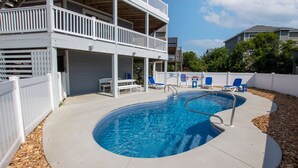 A swimming pool with a curved edge, surrounded by a white fence, and a covered patio area with blue chairs and a table. - Oceanside, Dogs Welcome, Private Pool, 800 ft to Beach Access, OS2E, Simply A-Mazzing (Corolla)