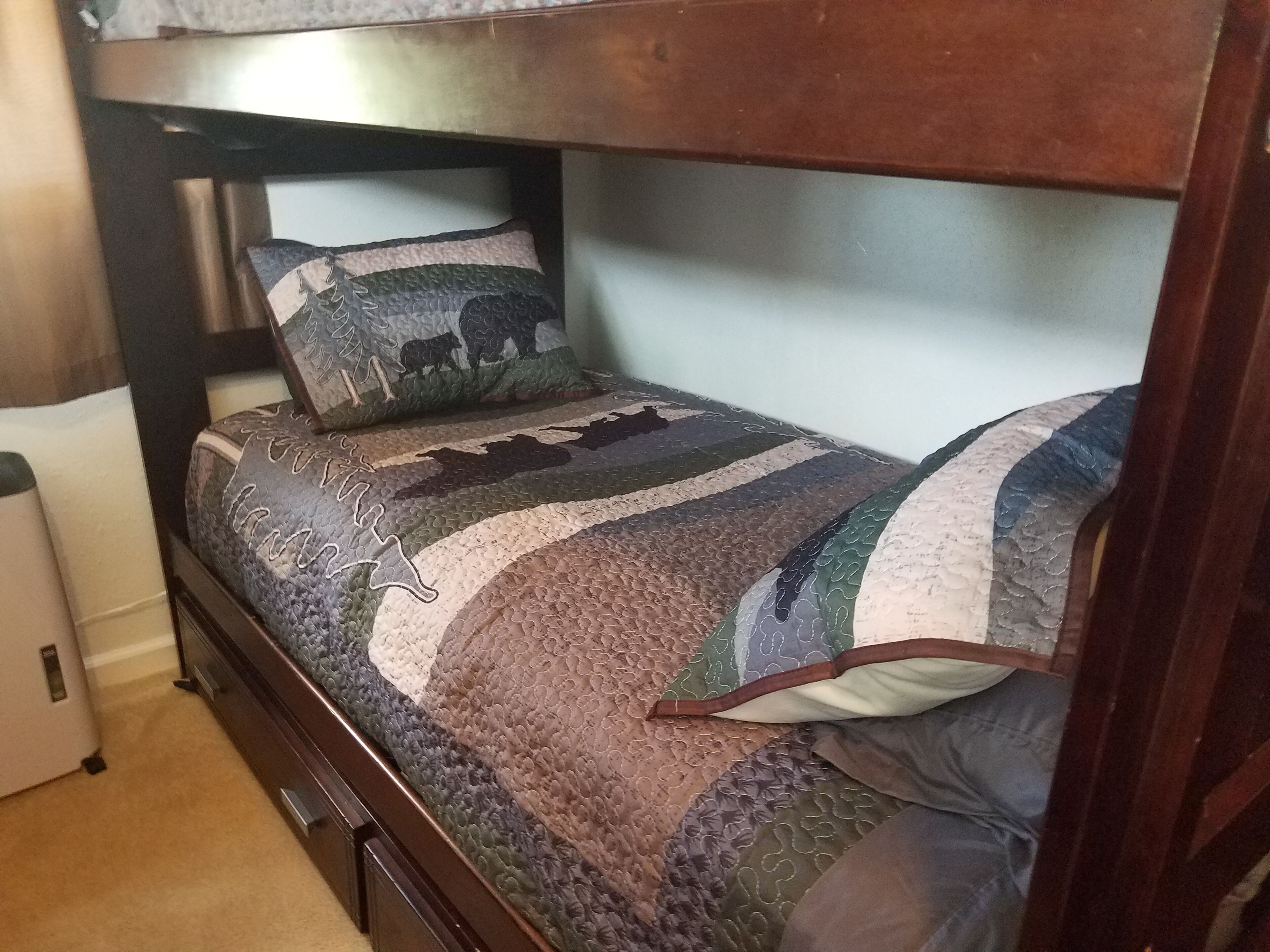 A bunk bed with a patterned quilt and pillows, a wooden headboard, and a small bedside drawer.