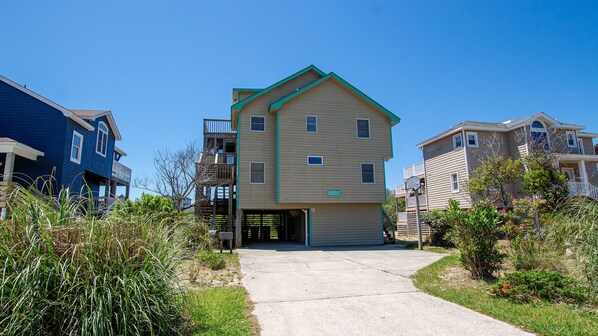A multi-story residential building with a central garage entrance, surrounded by greenery and adjacent houses. - Oceanside, Hot Tub, Close to Beach, Community Pool, CP4, Crown Palace (Corolla)