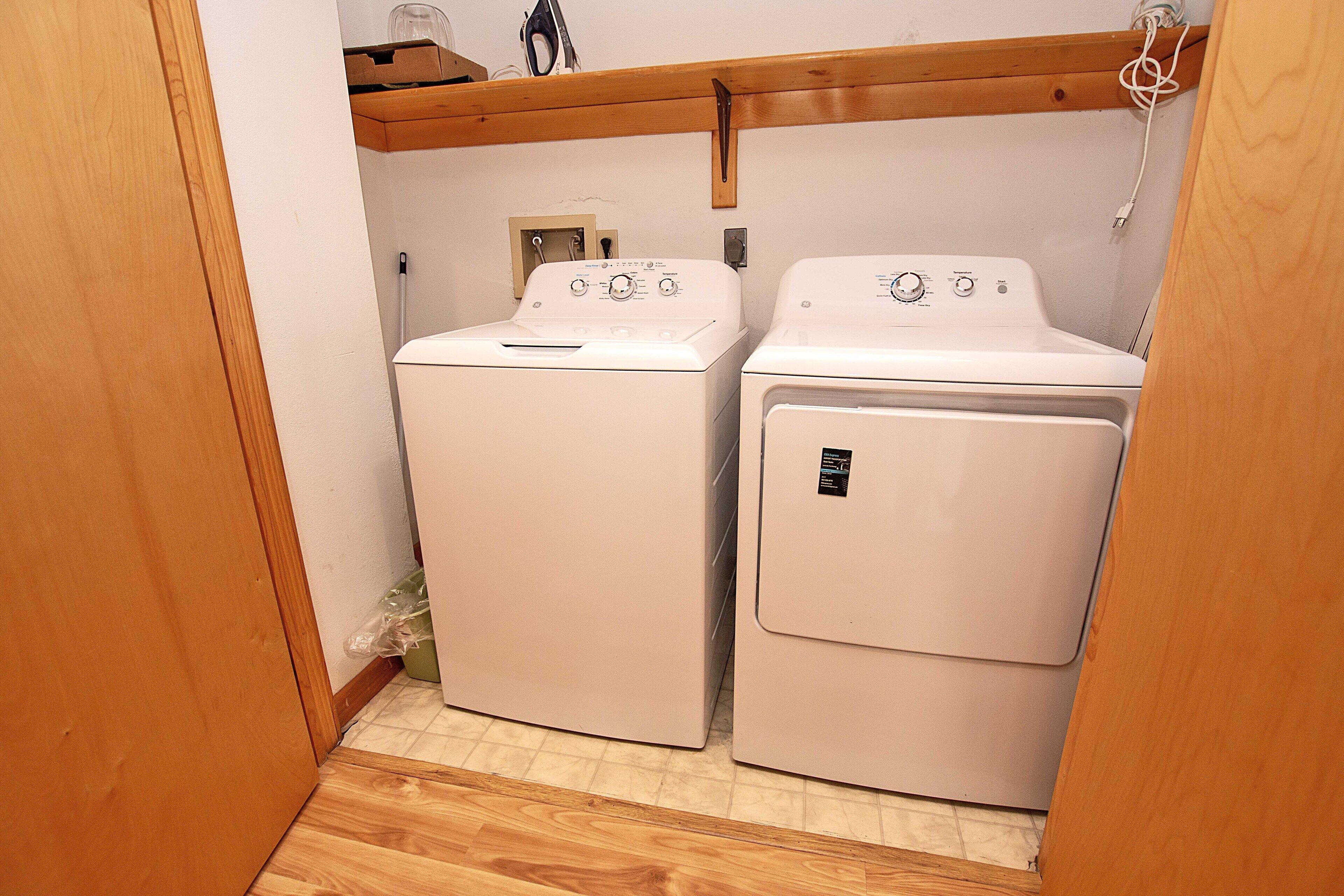 A laundry room with two white washing machines and a wooden shelf above.