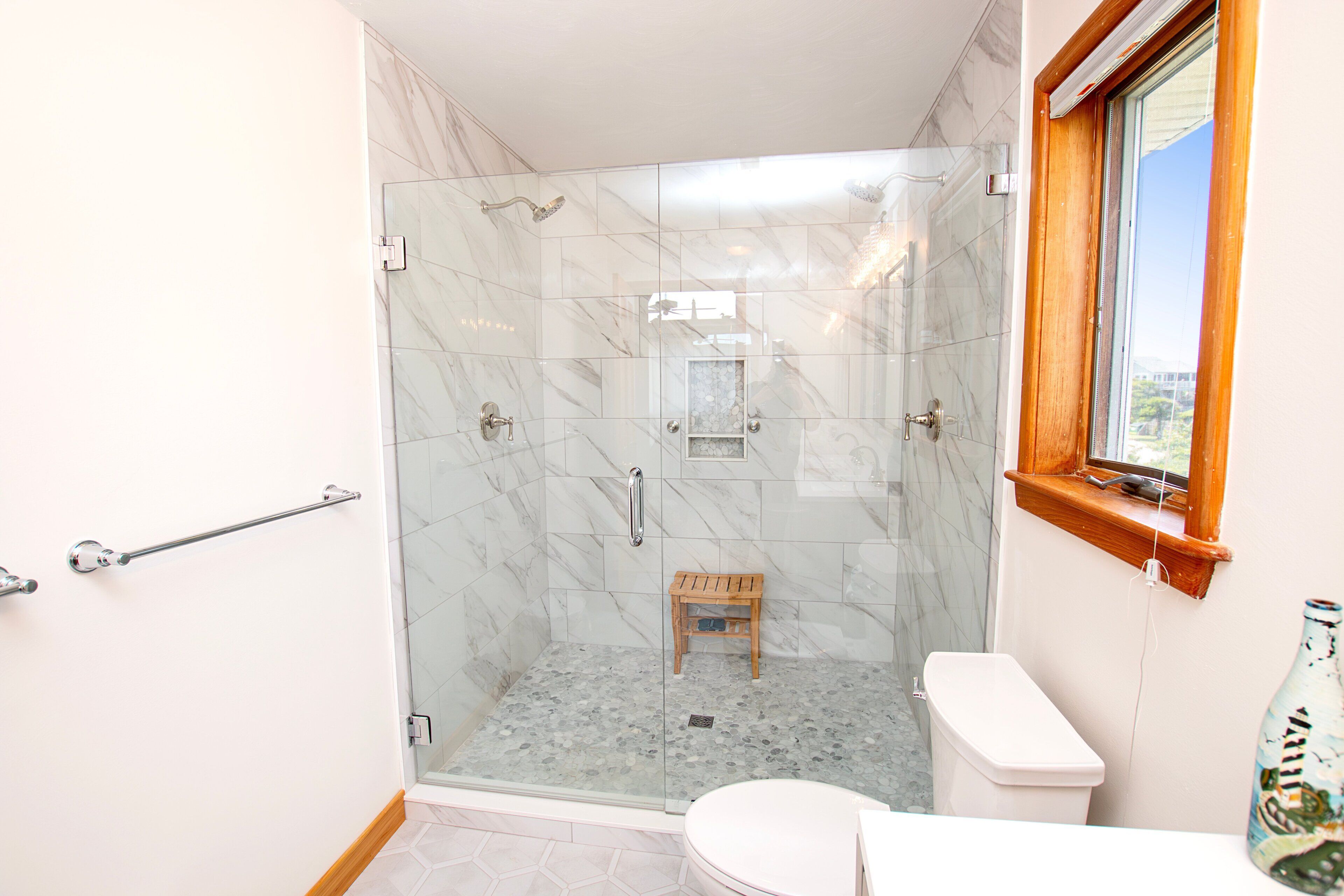 Une salle de bain avec une douche à parois de verre, un tabouret en bois, des toilettes blanches et une fenêtre avec vue.