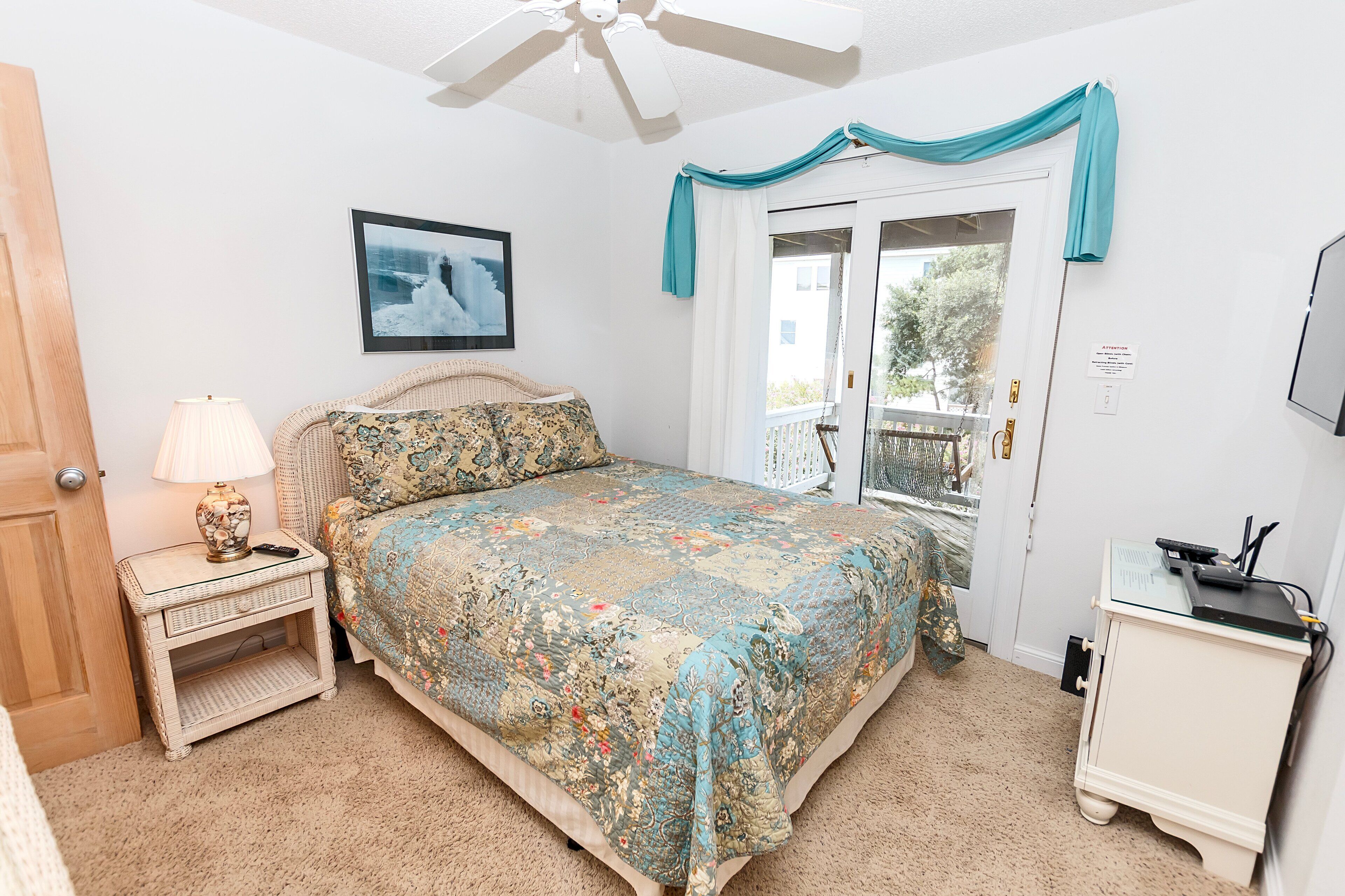 A bedroom with a bed, a nightstand, a lamp, a ceiling fan, a window with curtains, and a door.