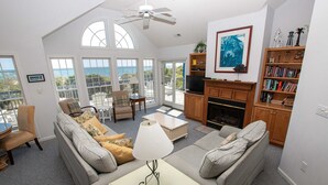 A spacious living room with a sofa set, a coffee table, and a television. There are large windows offering a view of the ocean.