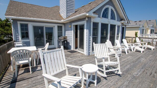 A wooden deck with white Adirondack chairs and a table, overlooking a beach.