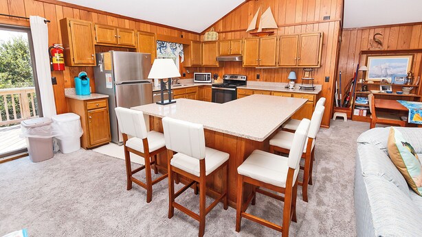 A kitchen with wooden cabinets, a central island with chairs, a refrigerator, microwave, stove, and a dining area with a table and chairs. - Oceanside, Private Pool, Hot Tub, Screened Porch, WC940, Whalin (Corolla)