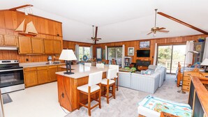 A spacious kitchen with wooden cabinets, a central island with bar stools, a dining area with a sofa, and a balcony with a view. - Oceanside, Private Pool, Hot Tub, Screened Porch, WC940, Whalin (Corolla)