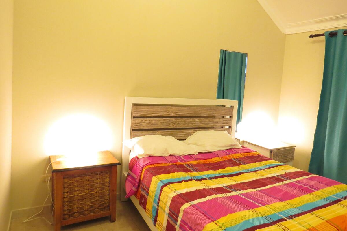 Une chambre à coucher comprenant un lit, deux tables de chevet, une commode, une tête de lit en bois et une fenêtre avec des rideaux.