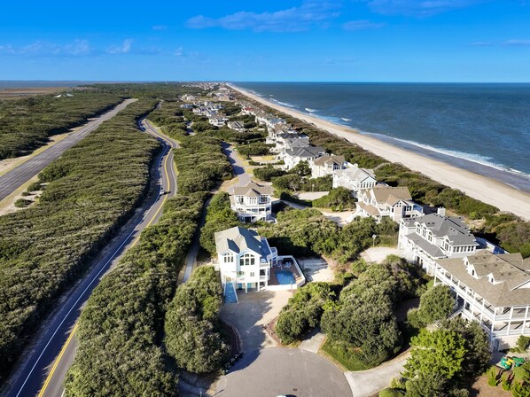 An aerial view of a coastal residential area with a beachfront, a road curving through the landscape, and houses with varying architectural styles.