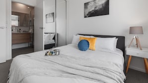 A modern bedroom with a bed, a bedside table, a lamp, and a painting on the wall. - Heart of Ormond Apartment  by Ready Set Host (Ormond)
