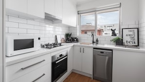 A modern kitchen with white cabinetry, a microwave, oven, and dishwasher. There is a window with a view of buildings outside. - Heart of Ormond Apartment  by Ready Set Host (Ormond)