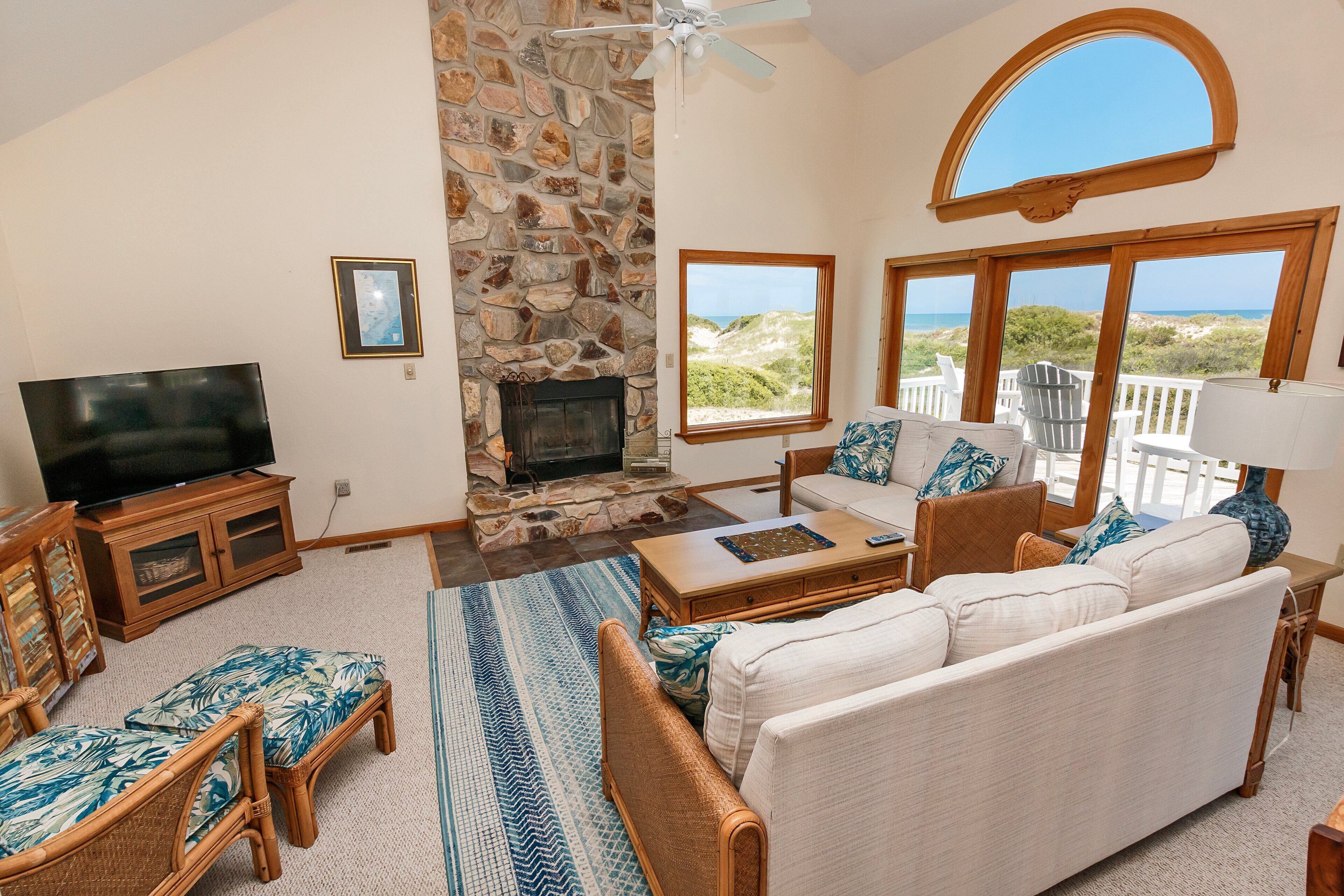 A living room with a stone fireplace, a television, wicker furniture, and large windows overlooking the beach.