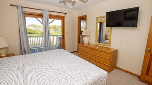 Une chambre à coucher avec un lit, une commode, un téléviseur, une fenêtre avec des rideaux et une vue sur la verdure à l’extérieur.