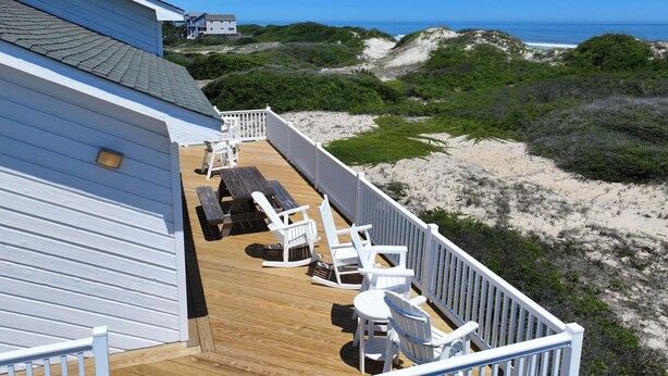 Une terrasse en bois avec des chaises blanches et une table, donnant sur une plage bordée de dunes et sur l’océan.