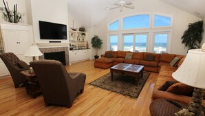 A spacious living room with a fireplace, a large window with a view of the ocean, and a cozy seating area. - OCEANFRONT, 8 BRs, Rec Rm, Pool, H Tub, Media Rm, Ocean Views, OS24C, Flying Cloud II (Corolla)