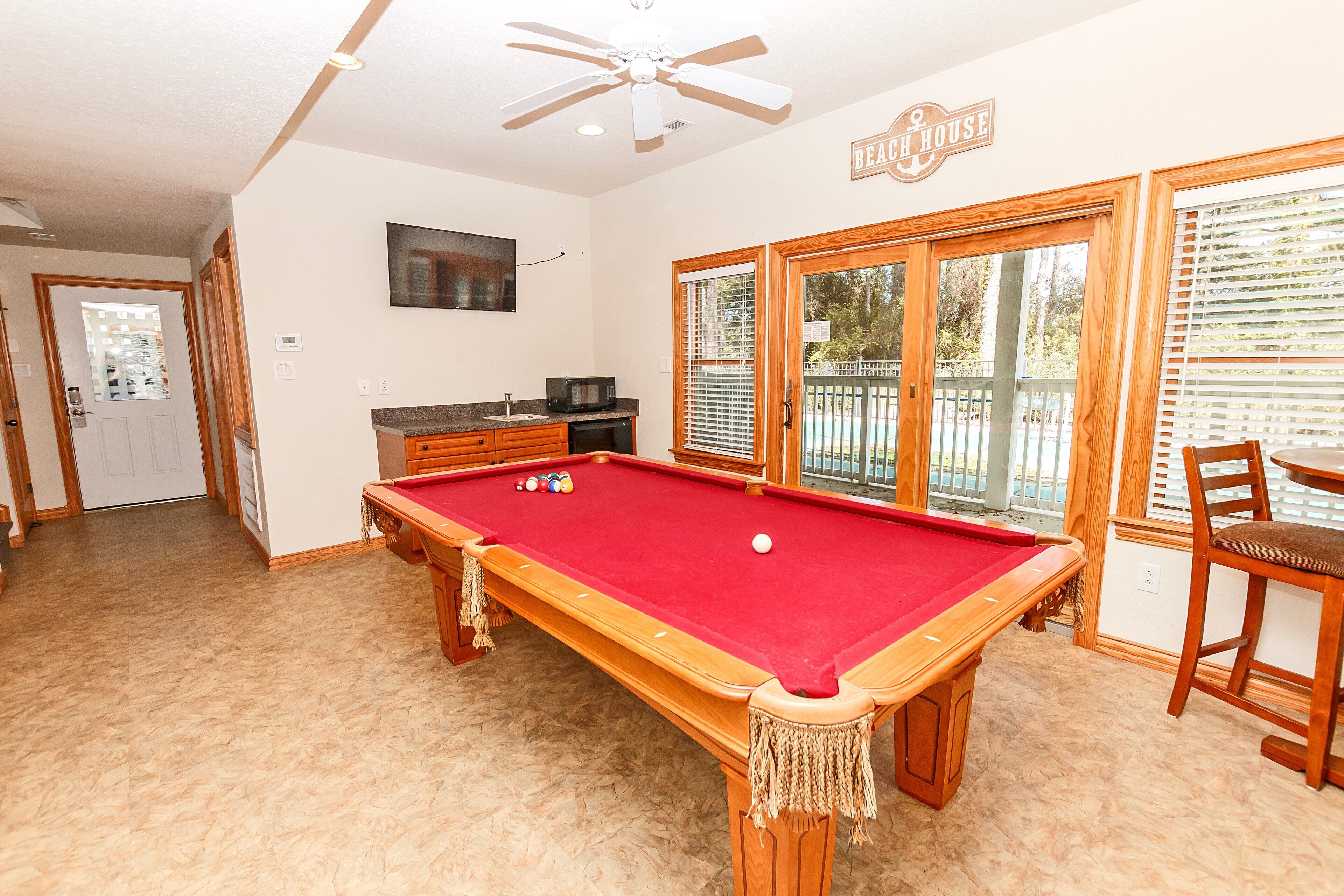 Een pooltafel met een rode viltbekleding, een plafondventilator, en een bord met de tekst 'Beach House'.