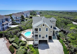 A large house with a swimming pool, surrounded by lush greenery and a view of the ocean. - Semi-Oceanfront, Rec Rm, Pool, Hot Tub, Close to Beach! PI191, OBXcape (Corolla)