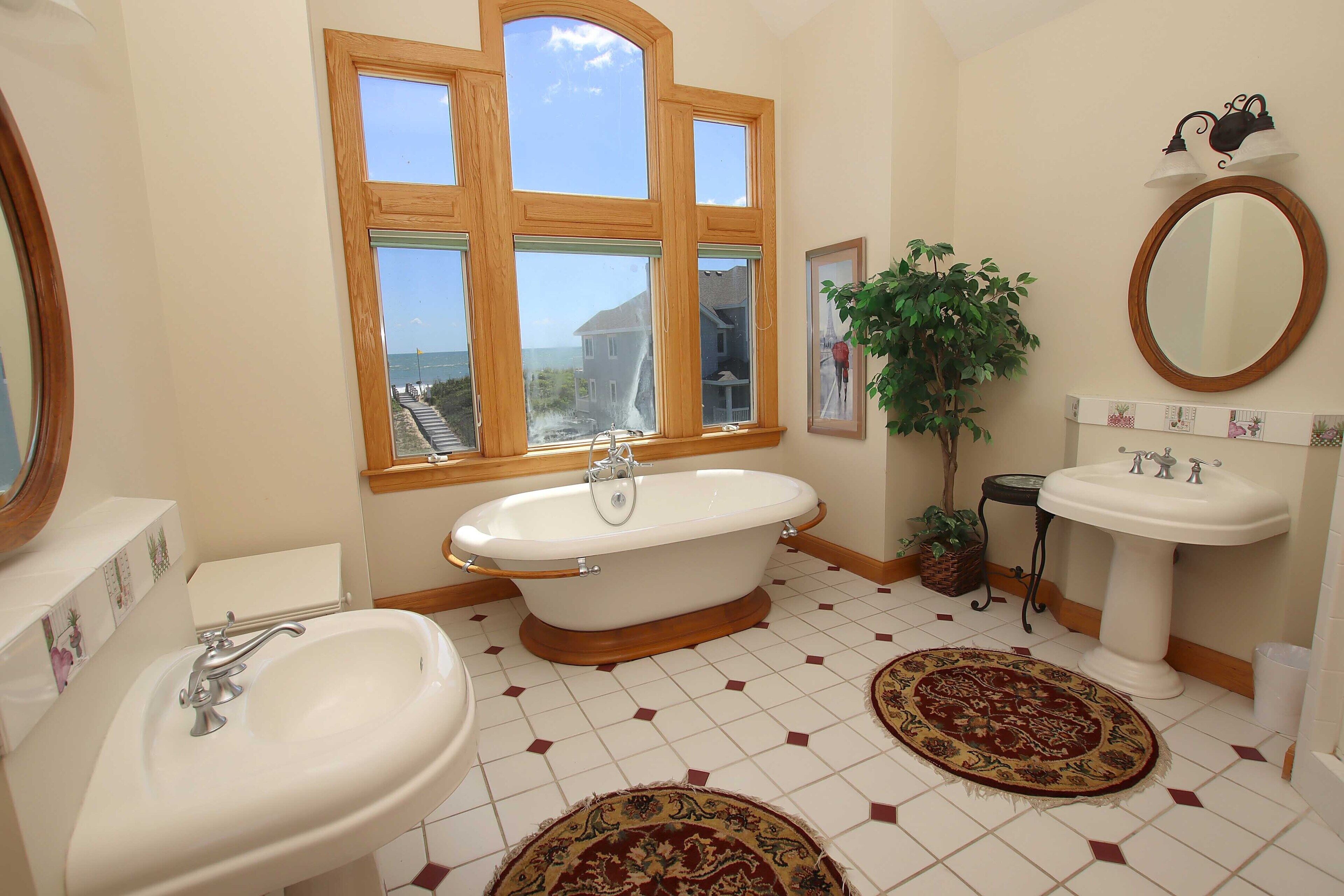 A bathroom with a bathtub, two sinks, a large mirror, a window with a view, and decorative rugs.