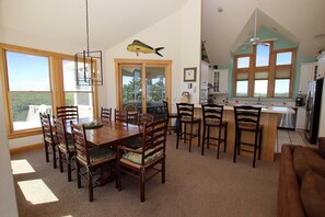 A spacious dining area with a large wooden table and chairs, a kitchen with a view, and a ceiling fan. - Semi-Oceanfront, Rec Rm, Pool, Hot Tub, Close to Beach! PI191, OBXcape (Corolla)