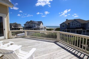 A wooden deck with white chairs, overlooking a beach with houses and a clear sky.