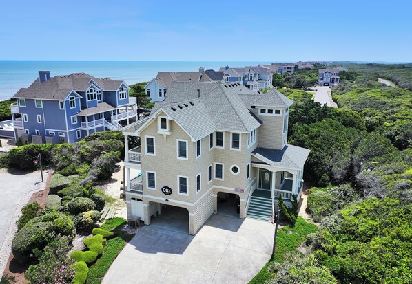 A large, multi-story house with a garage, surrounded by lush greenery and a view of the ocean. - Semi-Oceanfront, Rec Rm, Pool, Hot Tub, Close to Beach! PI191, OBXcape (Corolla)