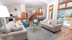 A spacious living room with a grey sofa, teal armchair, and a glass coffee table. A kitchen with white cabinets and a wooden staircase is visible in the background. - Semi-Oceanfront, Close to Beach, Private Pool, Rec Room, PI13, Ocean's 13 (Corolla)
