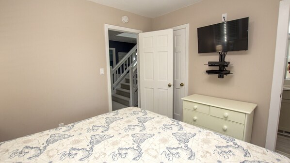 Une chambre à coucher comprenant un lit, une commode, un téléviseur fixé au mur et une porte donnant sur un escalier.