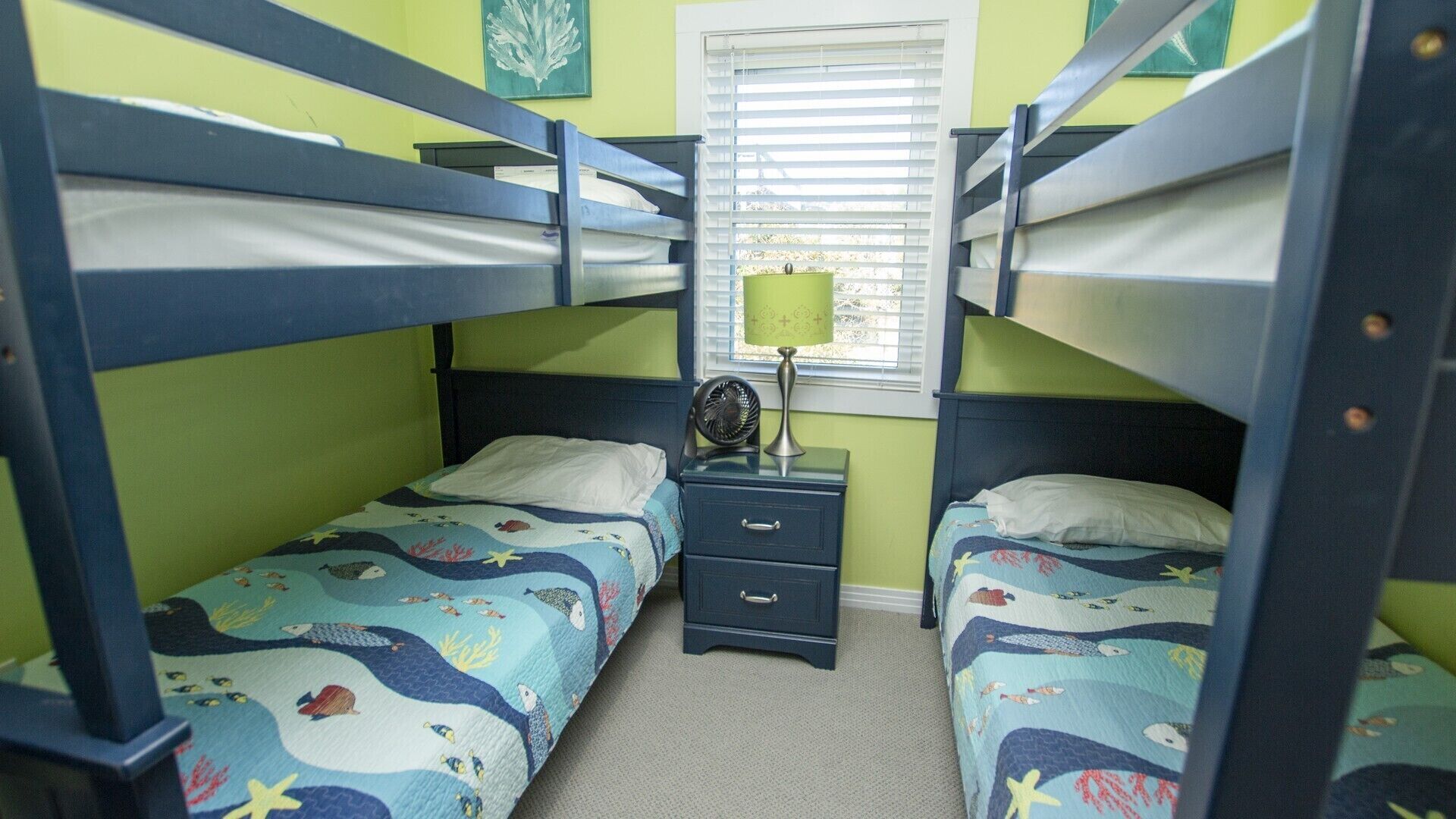 Dormitorio con literas, dos camas con cabeceras y pies de cama a juego. Entre las camas hay una mesita de noche con una lámpara y un ventilador. Las paredes son verdes y hay una ventana con persianas.
