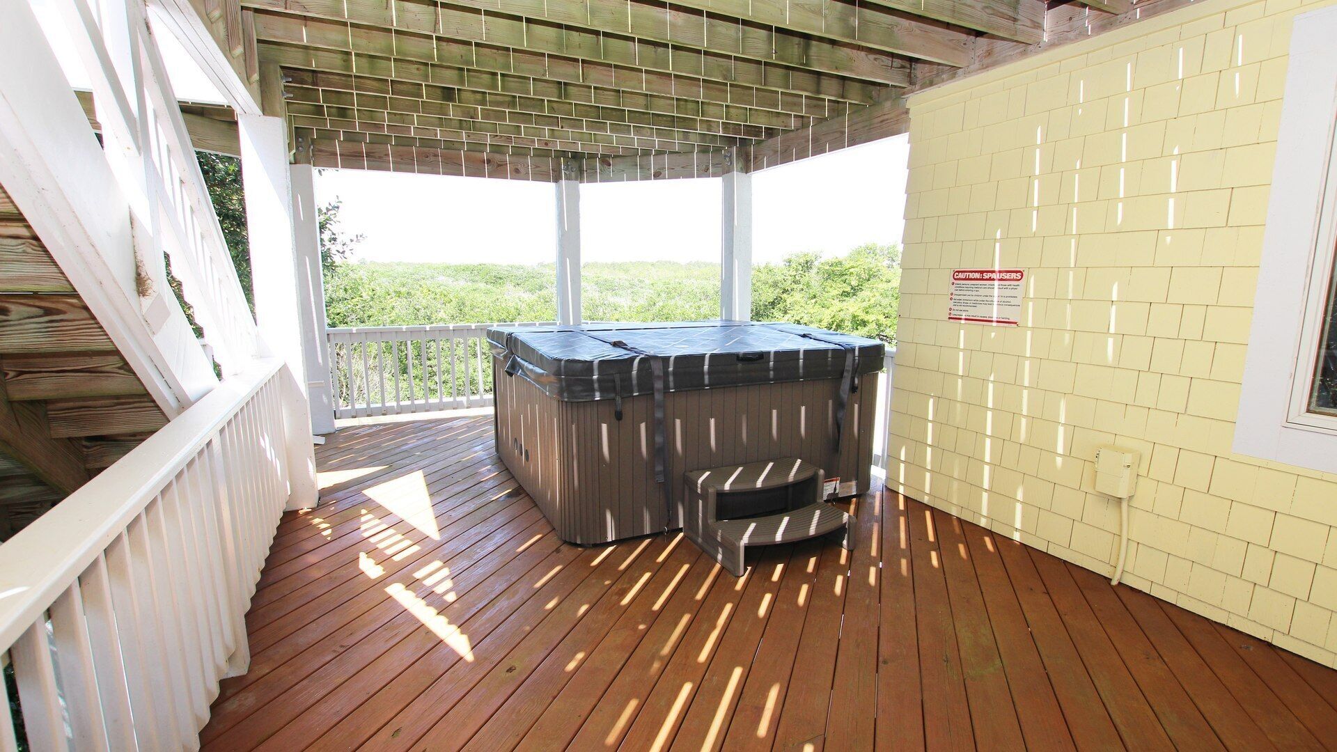 A covered outdoor deck with a hot tub, wooden flooring, and a white railing.