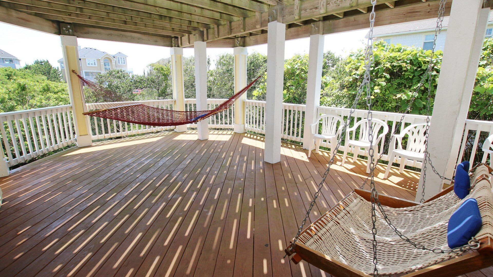A wooden deck with a hammock and a swing chair, surrounded by white railings and greenery.