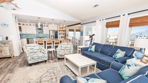 Ein modernes Wohnzimmer mit einem blauen Sofa, gemusterten Sessel und einem Couchtisch. Im Hintergrund ist eine Küche mit weißen Schränken und einem Essbereich zu sehen.
