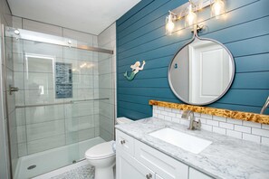 Un baño con ducha envuelta en vidrio, inodoro blanco, lavamanos de mármol y pared azul decorada con una sirena.