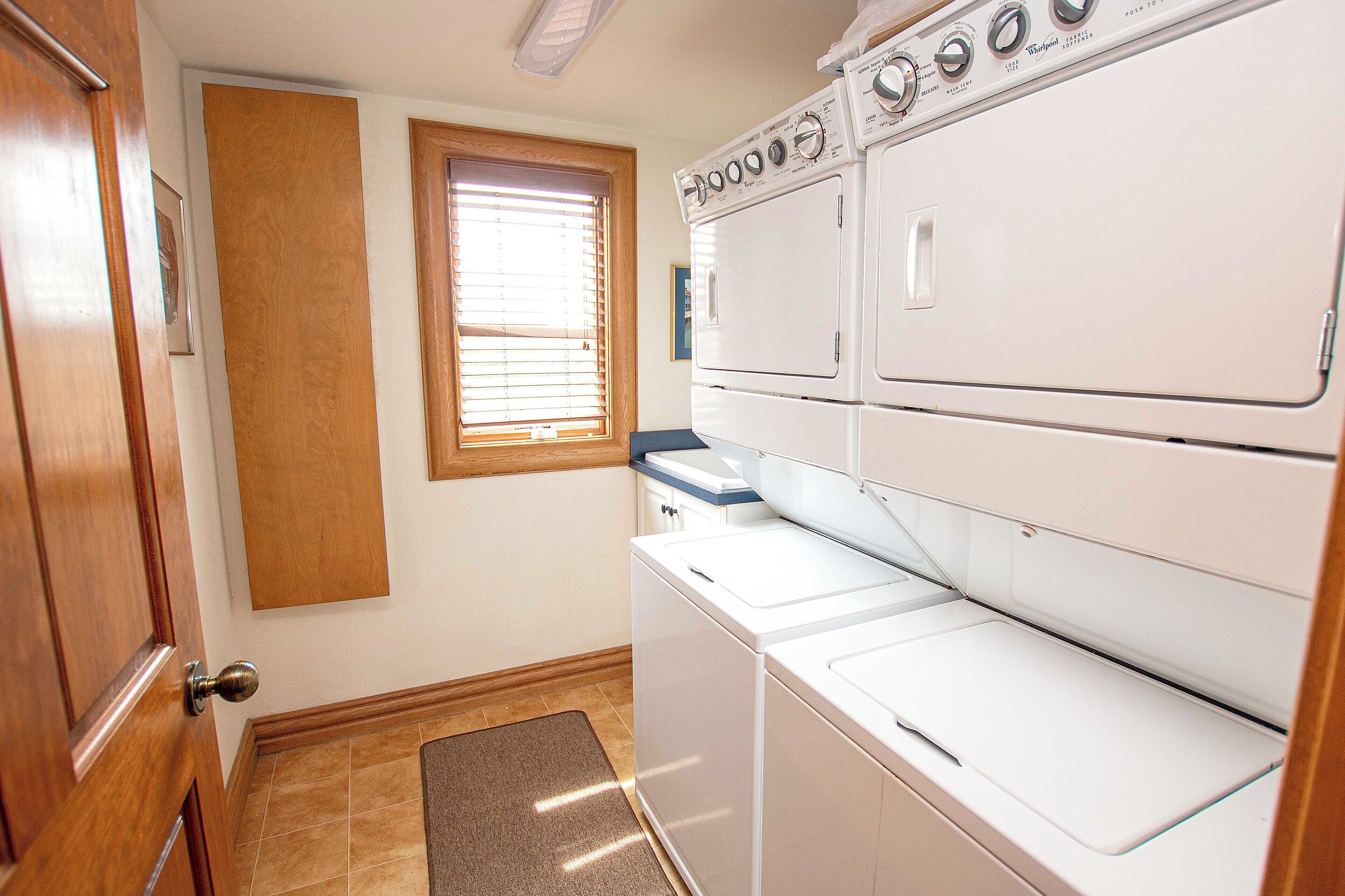 A laundry room with stacked washers and dryers, wooden doors, a window with blinds, and a brown mat at the entrance.